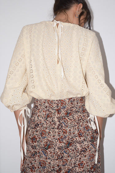 Person wearing a cream blouse with intricate patterns and a patterned skirt against a white background