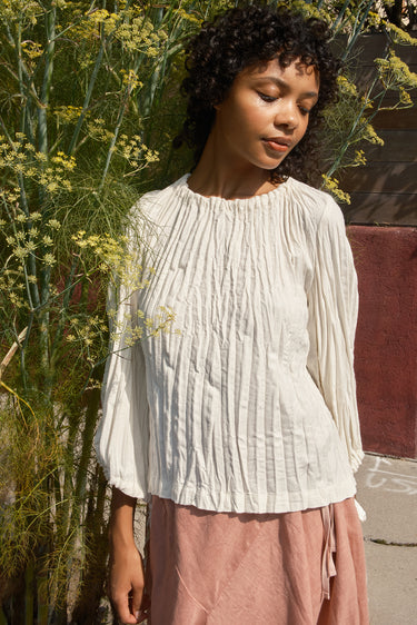 Woman wearing a white blouse and pink pants standing outdoors with greenery in the background