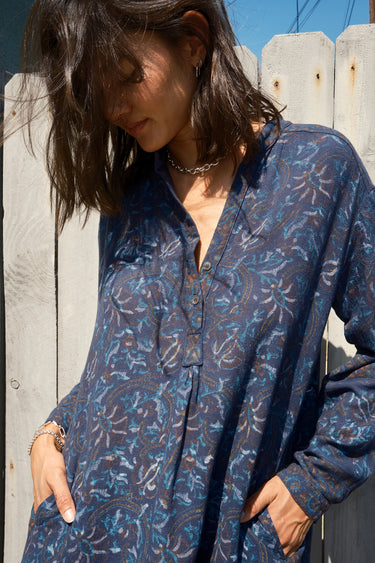 Woman standing in front of a grey fence wearing a navy blue tapestry mini shirt dress with long sleeves.