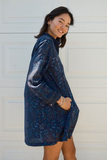 Side View of a woman standing in front of a white garage door wearing a navy blue tapestry mini shirt dress with long sleeves.