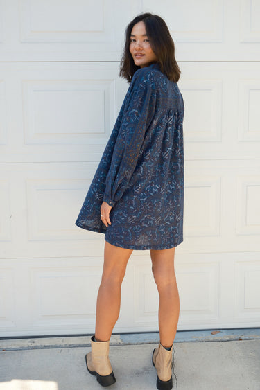 Back View of a woman standing in front of a white garage door wearing a navy blue tapestry mini shirt dress with long sleeves.