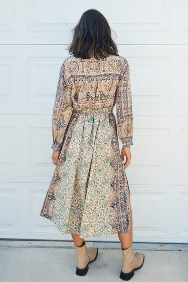 Woman  standing against a white garage door wearing a tan with green and black tapestry shirt caftan- dress, belted. 