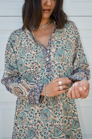 Woman  standing against a white garage door wearing a tan with green and black tapestry shirt caftan- dress. 