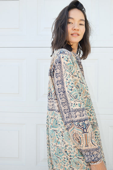 Woman  standing against a white garage door wearing a tan with green and black tapestry shirt caftan- dress. 