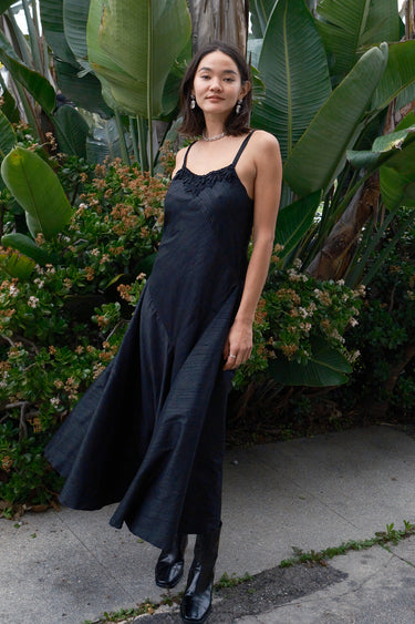 A woman standing on a pathway in a tropical setting, wearing a black, V-neck, sleeveless, maxi slip dress with darts on the side and adjustable straps.
