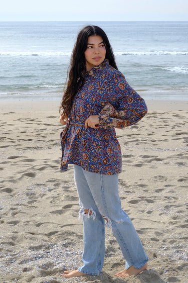 A woman wearing a blue a floral tapestry jacket with big patch pockets and an elastic shirred sleeve cuff, standing by the beach. She is also wearing jeans