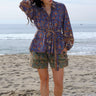 A woman wearing a blue floral tapestry jacket with big patch pockets and an elastic shirred sleeve cuff, standing by the beach. She is wearing coordinating shorts. 