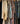 A close-up view of multiple patterned and solid long-sleeve garments hanging on wooden hangers from a clothing rack. The fabrics feature intricate paisley, floral, and textured prints in black, cream, teal, rust, blue, and green tones.