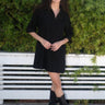 A woman standing in front of greenery wearing a black mini shirtdress with a collar, sleeves, and patch breast pockets, made of crinkle textured osnaburg cotton.
