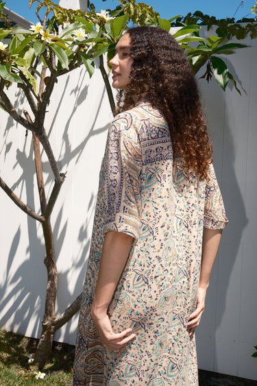 A woman standing outdoors wearing a floral print maxi dress with a shirt collar and sleeves, featuring a handloom Indian Tapestry fabric, back view.