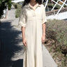 A woman standing on a pathway wearing an alabaster beige maxi shirtdress with a collar, sleeves, and a side slit, with a body made of crinkle textured osnaburg cotton.