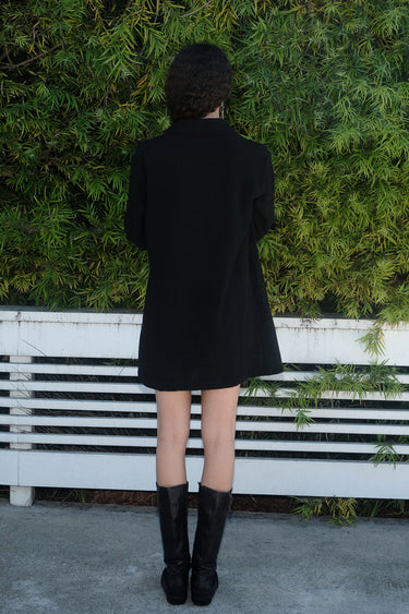 A woman standing in front of greenery wearing a black mini shirtdress made of crinkle textured osnaburg cotton, back view. 