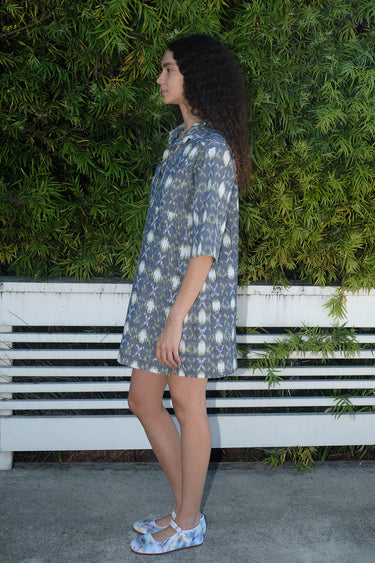 A person standing by a white fence with greenery wearing a mini shirt dress with a shirt collar and an "xo" Ikat pattern, side view.