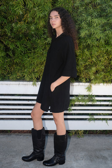 A woman standing in front of greenery wearing a black mini shirtdress with a collar, sleeves, and patch breast pockets, made of crinkle textured osnaburg cotton.