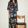 A woman inside standing next to a wooden chair, wearing a shirt dress caftan cut from a black ground tapestry printed with rainbow sun, moon and stars. 