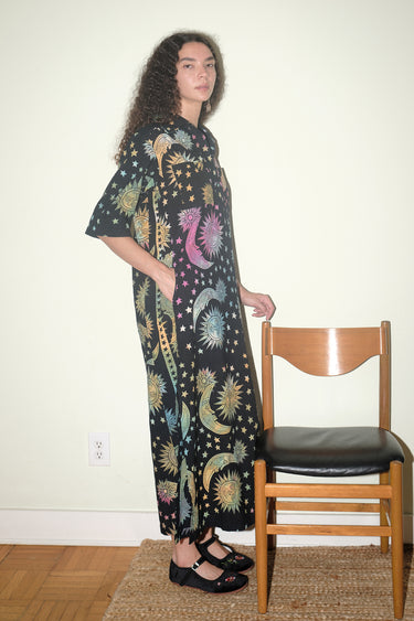 A woman inside standing next to a wooden chair, wearing a shirt dress caftan cut from a black ground tapestry printed with rainbow sun, moon and stars. 