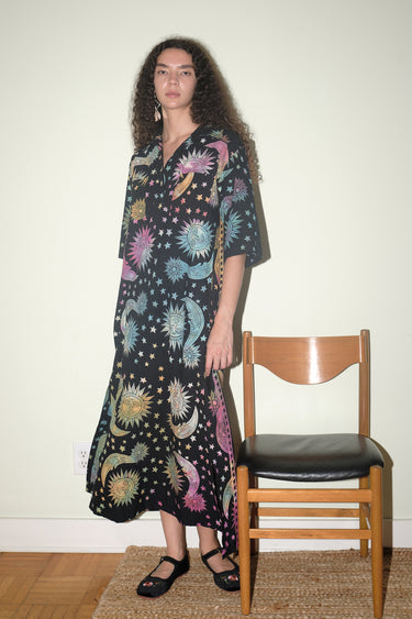 A woman inside standing next to a wooden chair, wearing a shirt dress caftan cut from a black ground tapestry printed with rainbow sun, moon and stars. 