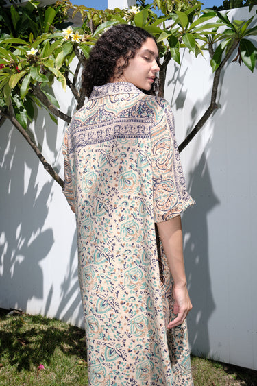 A woman standing outdoors wearing a floral print maxi dress with a shirt collar and sleeves, featuring a handloom Indian Tapestry fabric, back view.