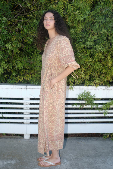 A woman standing outdoors wearing a blush calico tapestry maxi wrap dress with hand block printed tapestry design, side view.