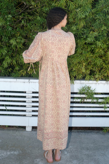 A woman standing outdoors wearing a blush calico tapestry maxi wrap dress with hand block printed tapestry design, back view.