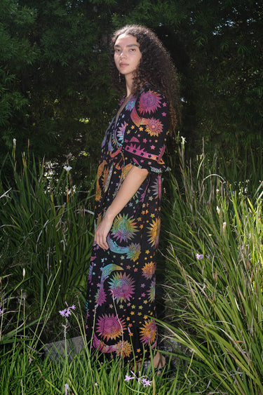 A woman standing in a garden wearing a multicolored maxi wrap dress with a tie-dye pattern and star shapes, featuring a wrap neckline and three-quarter sleeves.