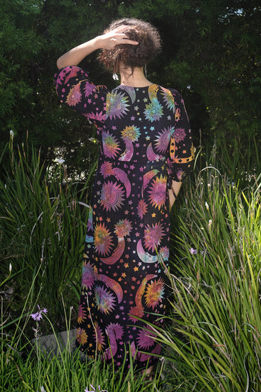 A woman standing in a garden wearing a multicolored maxi wrap dress with a tie-dye pattern and star shapes, featuring a wrap neckline and three-quarter sleeves, back view.