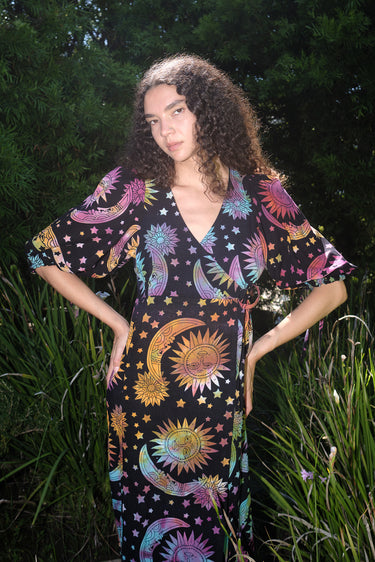 A woman standing in a garden wearing a multicolored maxi wrap dress with a tie-dye pattern and star shapes, featuring a wrap neckline and three-quarter sleeves.