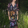 A woman standing in a garden wearing a multicolored maxi wrap dress with a tie-dye pattern and star shapes, featuring a wrap neckline and three-quarter sleeves.