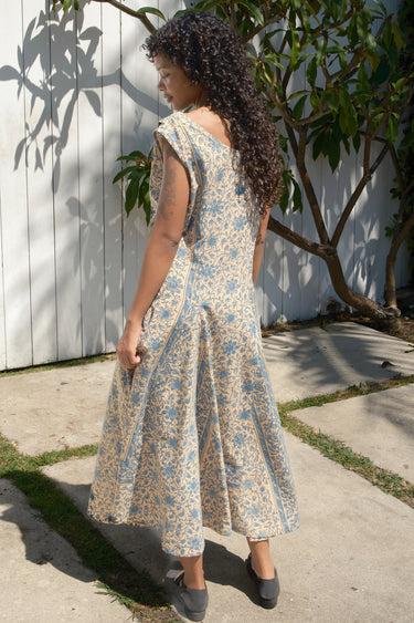 Woman wearing a floral dress standing outdoors near a white wall and tree.