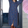 A woman wearing a  midnight sky handloom tapestry button front shirt dress with a band collar and side panels, featuring a tie at the back waist, standing in front of a door and a pink rose painted on a grey wall. 