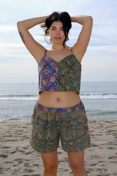 A woman standing at the beach wearing a floral tapestry low rise short cut using both green and blue tapestry of the same print tapestry mixed together features front pockets. Worn with a matching camisole. 