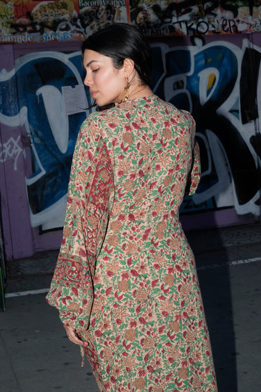 A woman wearing a maxi dress cut in a vintage rose handloom tapestry, featuring a wrap front and long full sleeves, standing in front of a graffiti-covered wall, back view.