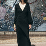 A woman standing in front of a colorful mosaic wall, wearing a black maxi dress with a wrap front, long full sleeves, and a bias quarter skirt.