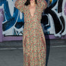 A woman wearing a maxi dress cut in a vintage rose handloom tapestry, featuring a wrap front and long full sleeves, standing in front of a graffiti-covered wall.
