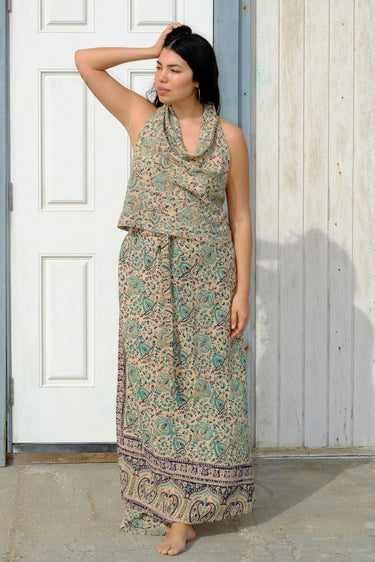 A woman standing by a building outside  wearing tan with green turquoise flower tapestry maxi wrap skirt and halter top. 