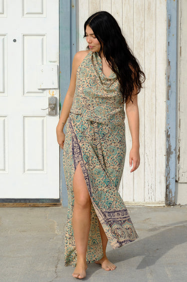 A woman standing by a building outside  wearing tan with green turquoise flower tapestry maxi wrap skirt and halter top. 