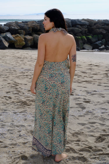 A woman standing at the beach wearing tan with green turquoise flower tapestry draped neck halter top with a matching skirt. Back View