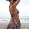 A person wearing sunglasses and a floral patterned woven swimsuit shorts, posing on a beach with the ocean in the background.