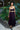 Woman in aNoire black  dress with yellow accents standing in front of green foliage.