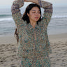 A woman standing at the beach wearing tan with green, turquoise flower tapestry shirt with bell sleeves and a matching skirt. 