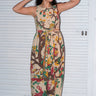 A woman wearing a beige maxi dress with a tree of life tapestry pattern, featuring wide ties and a slight empire waist. She is standing by a white roll up door.