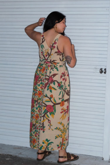 A woman wearing a beige maxi dress with a tree of life tapestry pattern, featuring wide ties and a slight empire waist. She is standing by a white roll up door. Back View