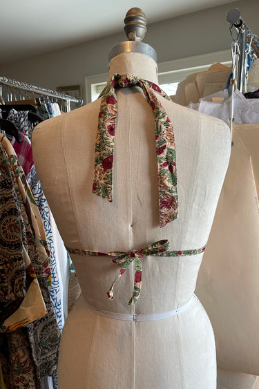 A halter style bikini top with a floral pattern in red and green on an ecru background, displayed on a dress form, back view.