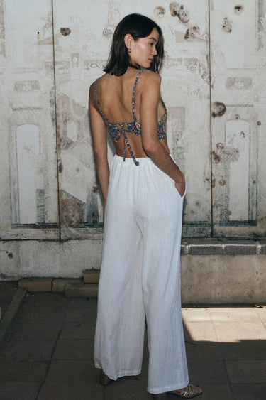 A woman standing by a mural wearing bianca bra top with white trousers. Back View.