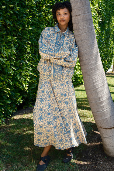 Woman in a floral jacket over a dress standing next to a palm tree in a garden setting