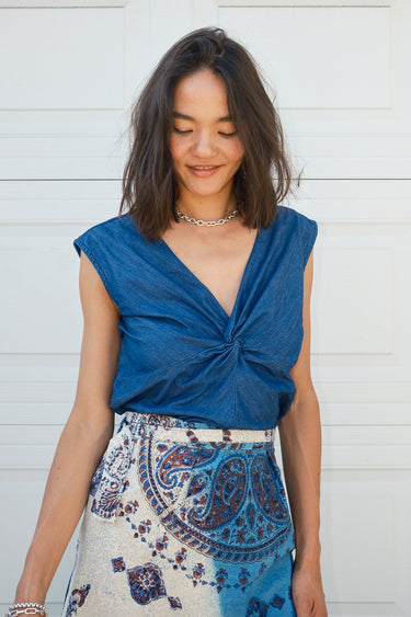 A woman standing  by a white garage wearing a denim top with a V-neck and shirred knot at the end of the neckline and a tapestry skirt.