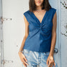 A woman standing against a door frame, wearing a light weight denim  top with a V-neck and shirred knot at the end, paired with blue jeans a
