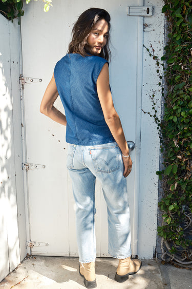 A woman standing against a door frame, wearing  denim top with a V-neck and shirred knot at the end, paired with blue jeans and tan ankle boots. Back View