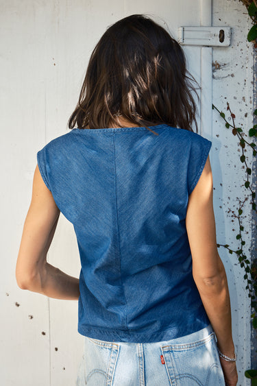 A woman standing against a door frame, wearing a  denim top with a V-neck and shirred knot at the end, paired with blue jeans.