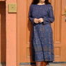 A woman standing in front of an orange  door, wearing a blue tapestry wrap dress with long bell sleeves and a quarter circle skirt, featuring patch pockets.